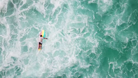 Aerial-drone-birds-eye-view-of-surfer-with-colorful-board-paddling-out-in-light-blue-water-in-Hawaii