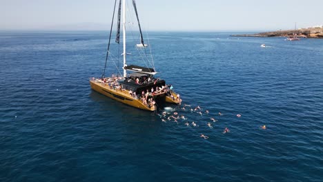 man-jumps-from-boat-into-water,-catamaran-in-blue-sea-water,-beautiful-bay-on-an-island,-tenerife,-boat,-summer,-drone