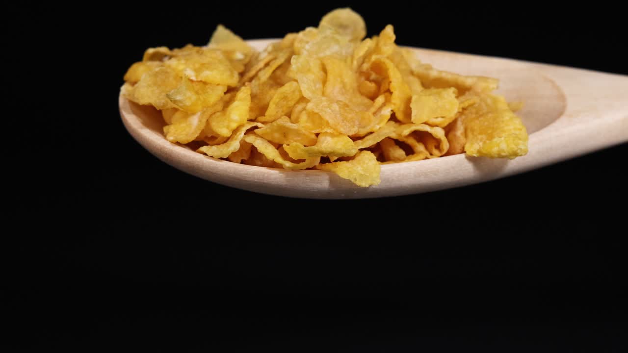 Cornflakes cascade from a wooden spoon, set against a dark backdrop, highlighting texture and motion in a controlled lighting environment