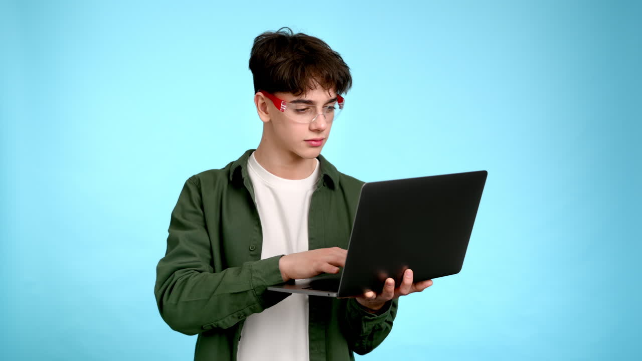 Young man in glasses using his laptop and smiling on a blue background