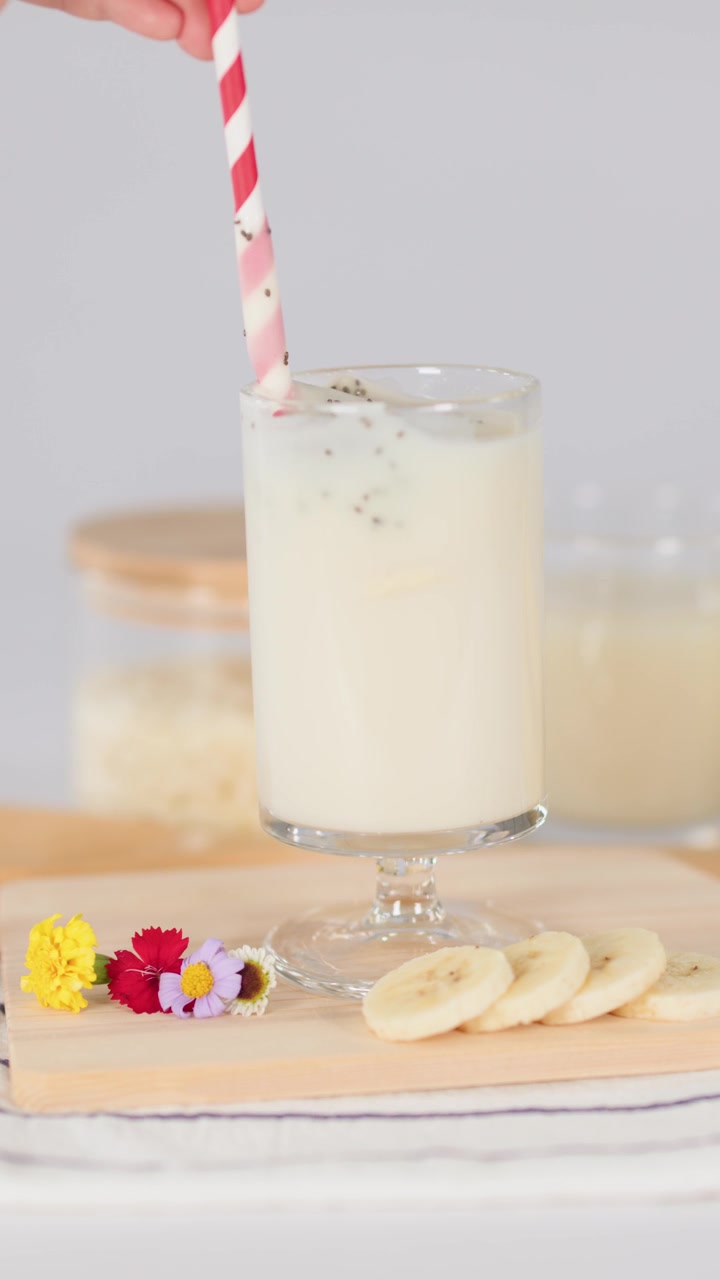 Una mano revuelve un batido cremoso de plátano en un vaso con una pajita a rayas sobre una mesa luminosa.