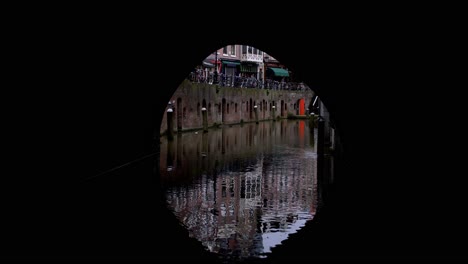 view from a boat peacefully cruising down a canal in utrecht, netherlands: moving through a dark tunnel under a bridge, seeing the cityscape reflecting in the water and panning up to the skyline