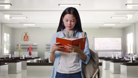 estudiante adolescente asiática con una mochila sosteniendo y leyendo un libro mientras está de pie en el laboratorio de ciencias