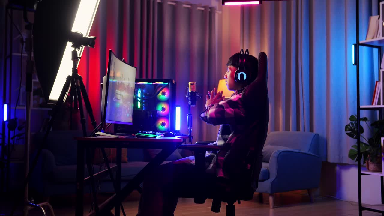 vista lateral de un streamer asiático que se estira mientras juega un juego a través de la red en una computadora personal. videojuego de transmisión en vivo, escritorio iluminado por luz de tira led rgb