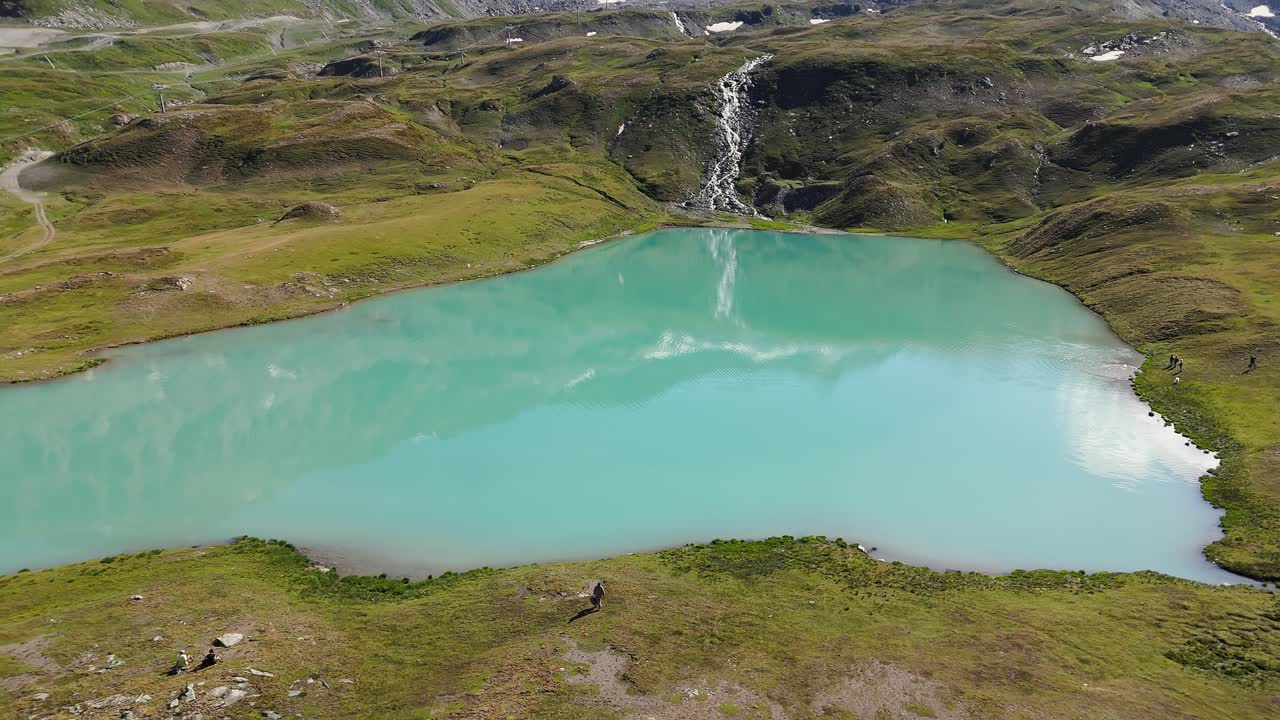 Aerial shot rising to reveal a stunning blue-green lake in Cervinia, Italy, with a traveler setting off on an adventure