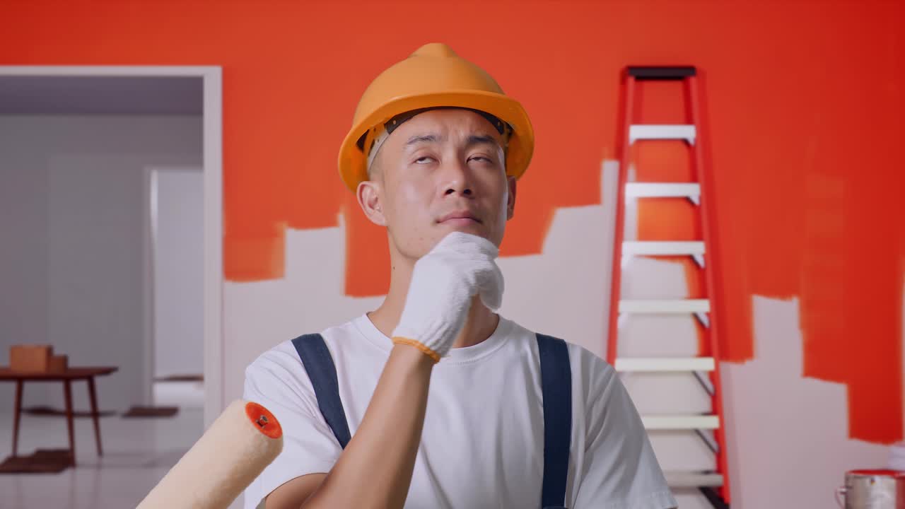 Close Up Of Asian Man Painter Wearing Safety Helmet Thinking And Looking Around Then Raising His Index Finger While Standing With Painting Wall In Room