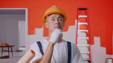 close up of asian man painter wearing safety helmet thinking and looking around then raising his index finger while standing with painting wall in room