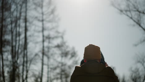 a father gently lowers his child from his shoulders, with a view of a bright sky and trees in the background