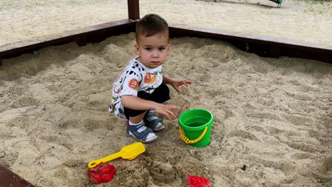 calm caucasian toddler sitting in the sandpit. baby boy puts his hands into the green bucket filled with water.