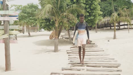 hombre afroamericano feliz llevando una tabla de surf caminando por el paseo marítimo en una playa soleada, cámara lenta