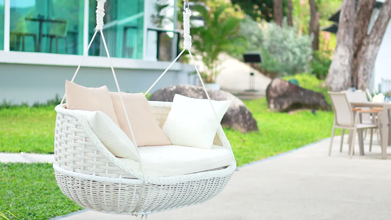 A white swing chair gently sways in a serene garden, surrounded by lush greenery and modern architecture in Phuket, Thailand