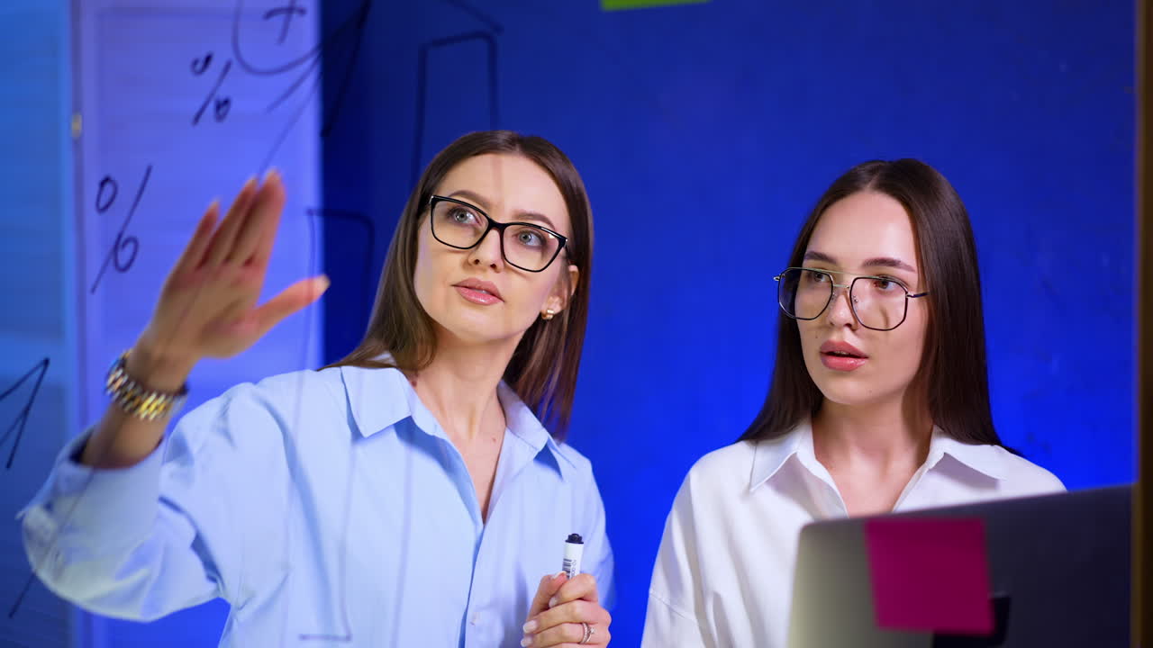 Two beautiful Caucasian ladies wearing glasses and white shirts stand at glass wall. Women talk, discuss, plan and look at laptop.