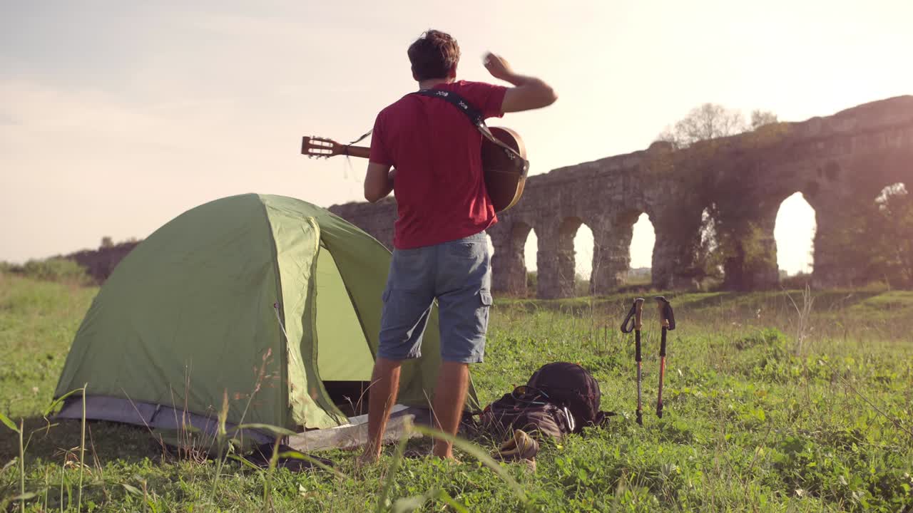 joven mochilero fuera de la tienda de campamento recoge la guitarra y camina por la hierba frente al acueducto romano arcos en el parco degli aquedotti ruinas del parque en roma al atardecer palos en el suelo en cámara lenta
