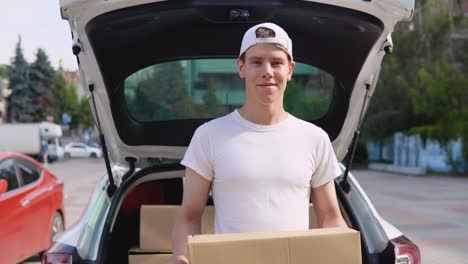 an employee holds a box in his hands and stands next to a car filled with parcels. moving and delivery of manufactured goods