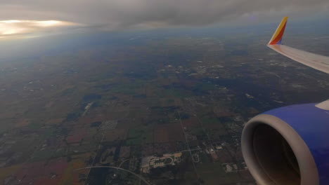 Shot-of-an-airplane-and-its-engine-and-wing-flying-in-top-of-a-city-with-the-clouds-above-as-the-sun-is-starting-to-set