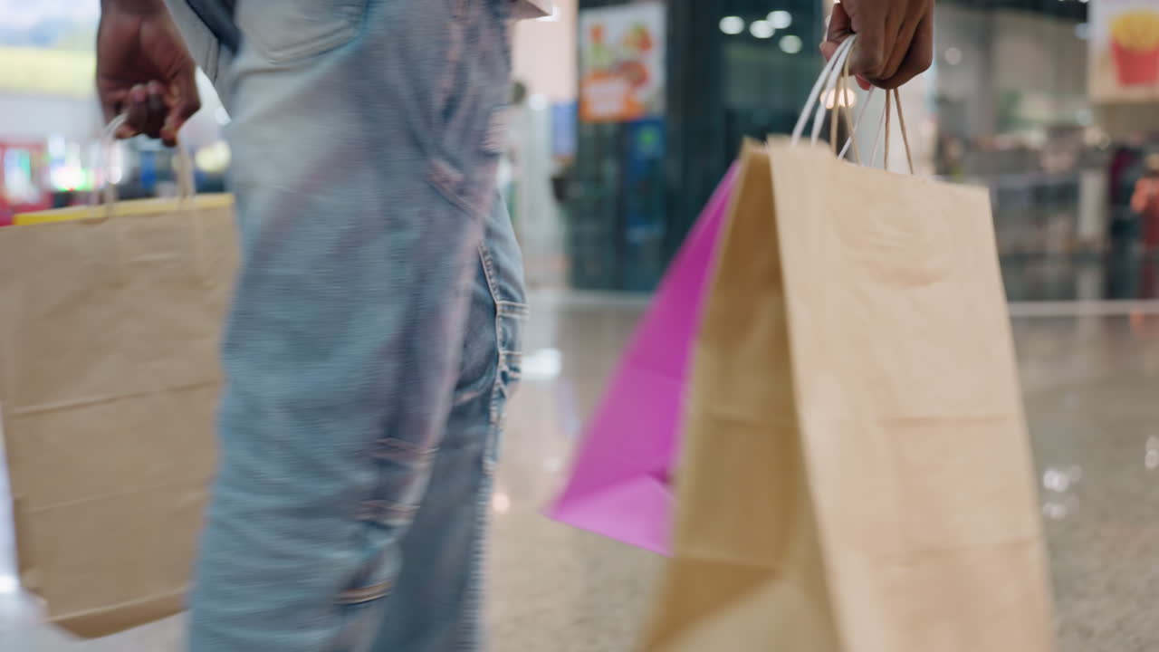 Close up of man carrying paper shopping bags in both hands while walking through shiny indoor shopping center with bright reflections on polished floor creating modern