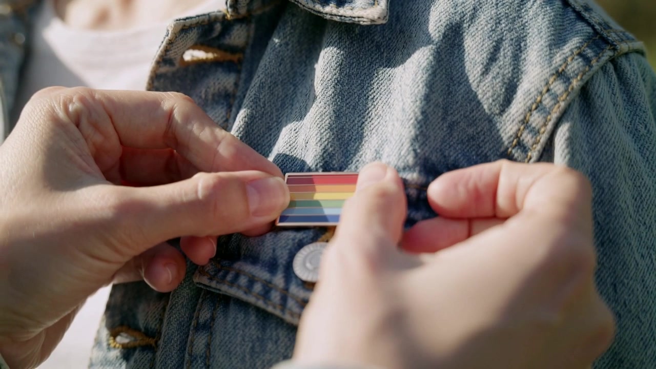 Approaching hands fastening rainbow-striped pin on denim jacket outdoors, showing support