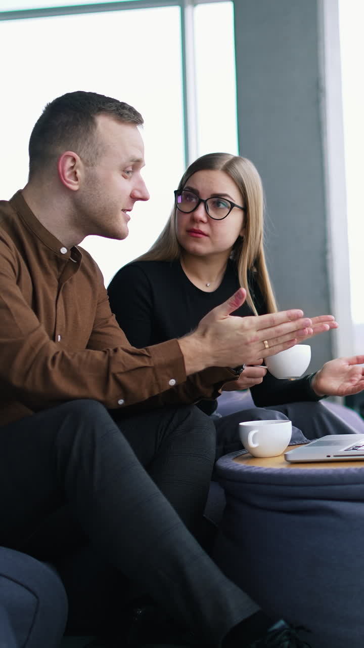 Young business people talk in modern office. Handsome businessman and female partner sitting in comfortable chairs and having a conversation in front of a laptop. Vertical video