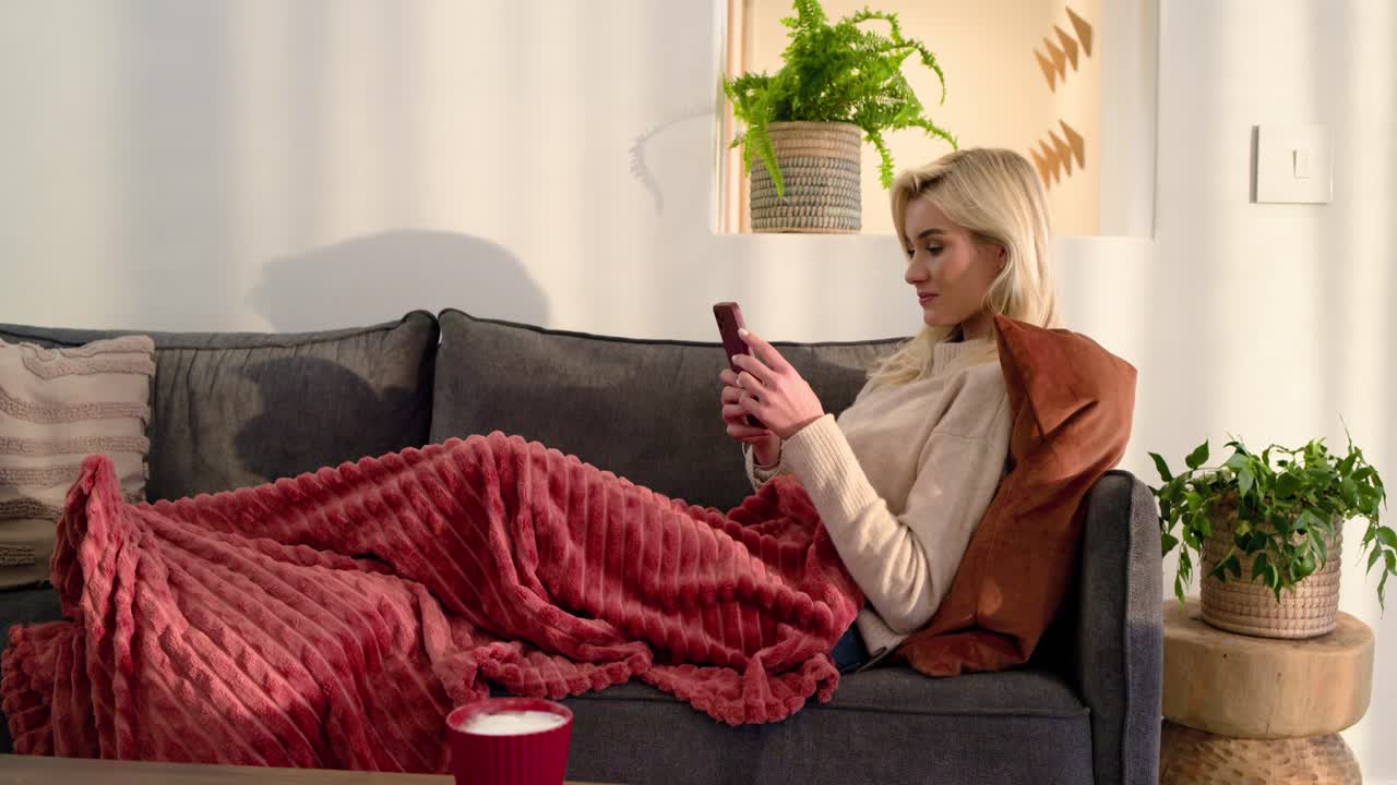 Female resting head, scrolling red phone on red-blanketed sofa, smiling, speaking, copy space