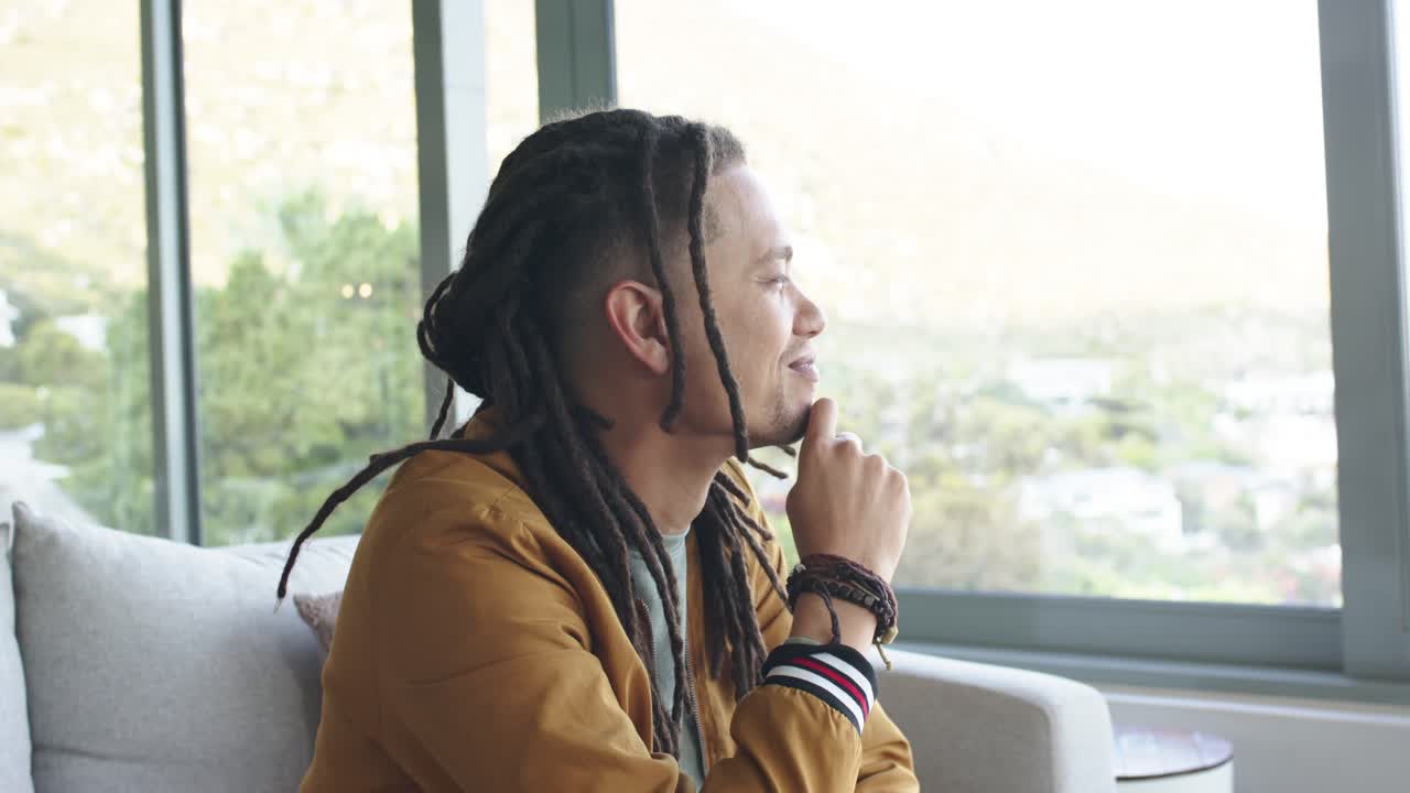 Hillside view inspiring African American man on sofa by window shifting gaze, smoothing dreadlocks