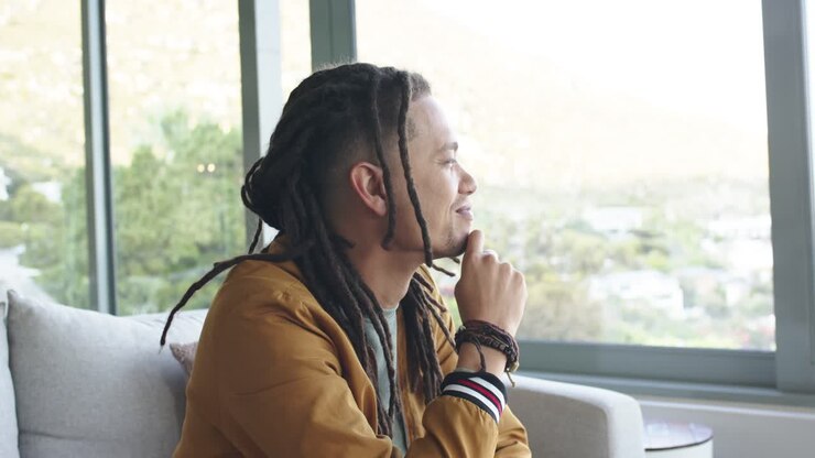 Hillside view inspiring African American man on sofa by window shifting gaze, smoothing dreadlocks