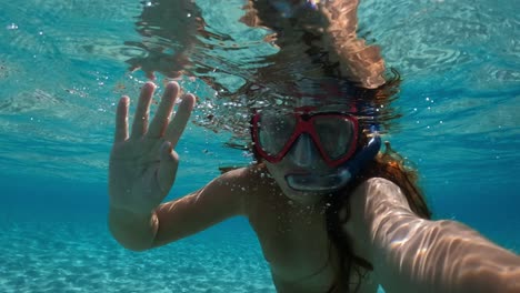 underwater selfie of little red-haired girl with diving mask and snorkel greeting waving hand and swimming in clear sea water