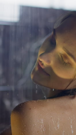 young tender blonde woman washes shoulder with hand under flowing water in shower booth against dark marble tile daily routine slow motion