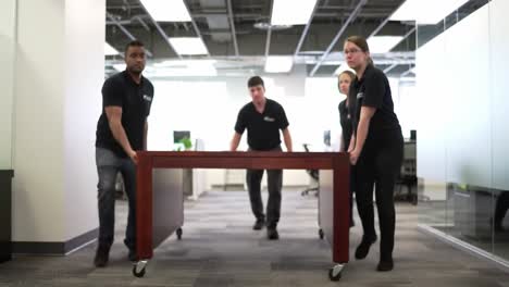 teamwork in motion: four individuals collaborate to move a heavy table through an office space, demonstrating cooperation and efficiency