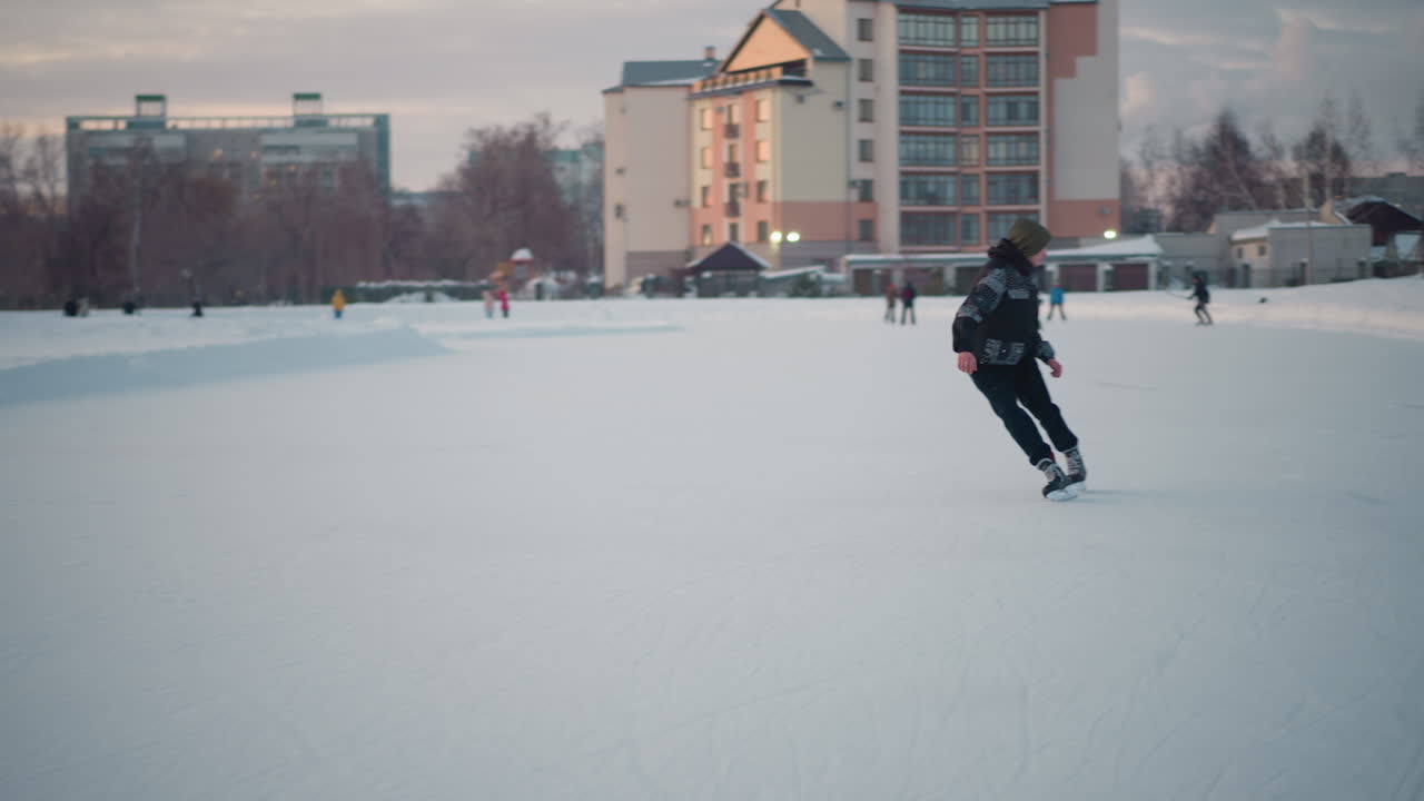 student skating backward on outdoor ice rink near urban buildings and snow, wearing patterned jacket and green hoodie, gliding smoothly past blurred skaters under pastel evening sky