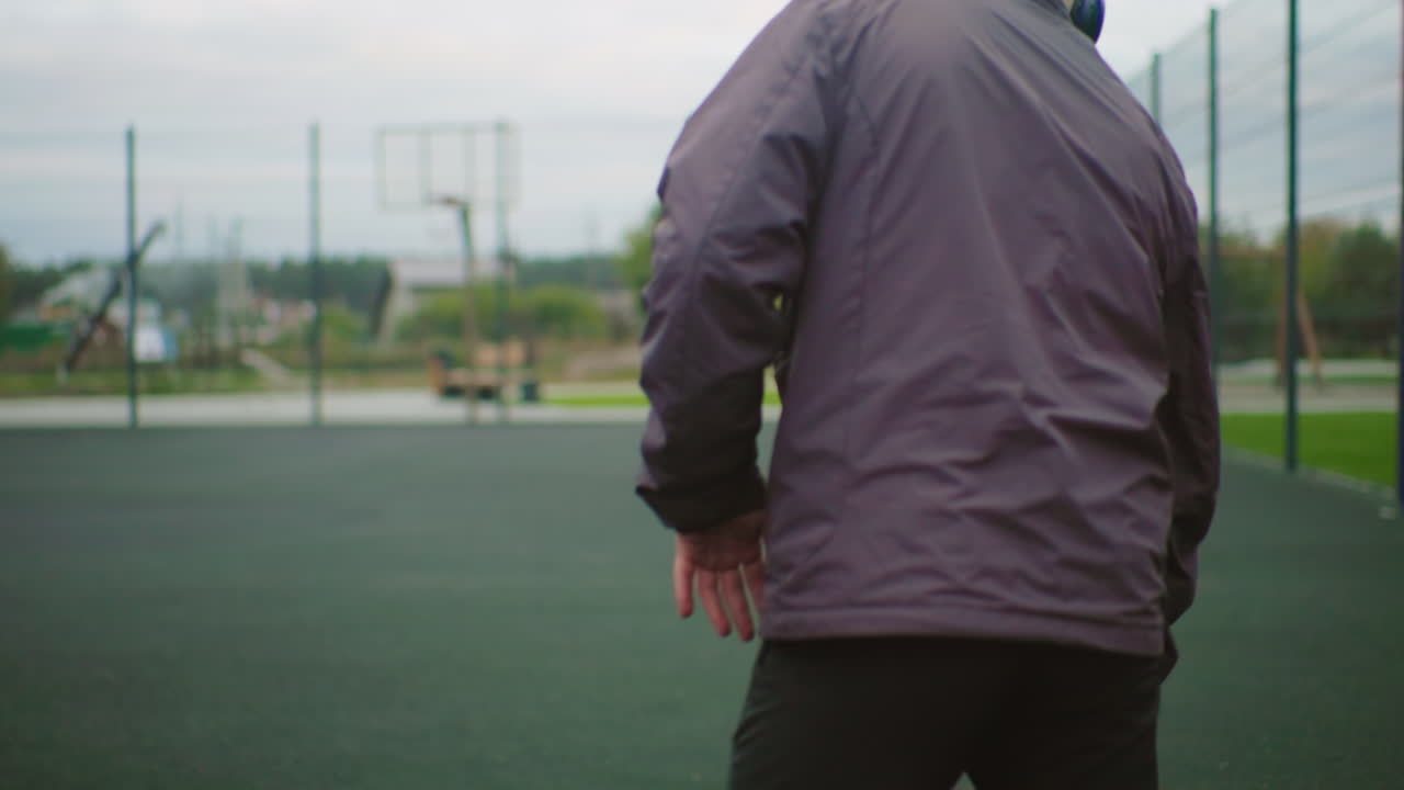 Male practicing dribbling on outdoor court wearing jacket beanie headphones, closeups of feet legs and back, slowmotion dribble on fenced asphalt under overcast sky, focused solitary training mood
