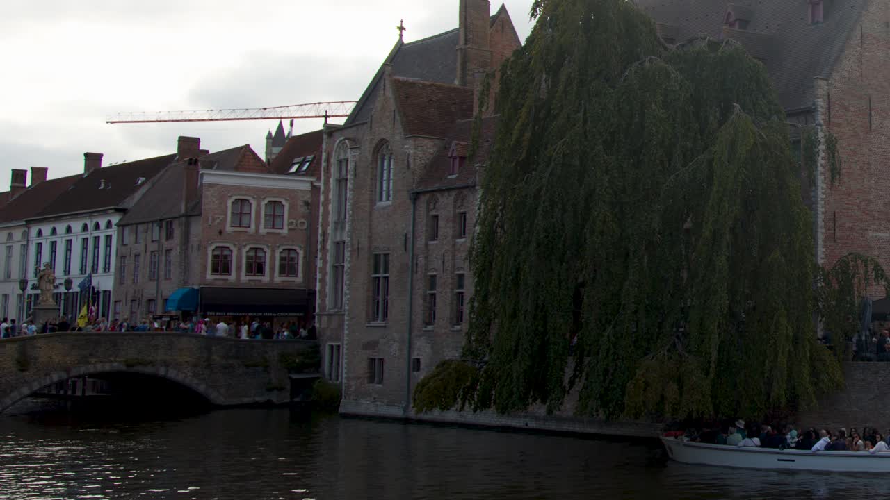 Tour boat glides past historic architecture and willow tree on Bruges canal, overcast daylight
