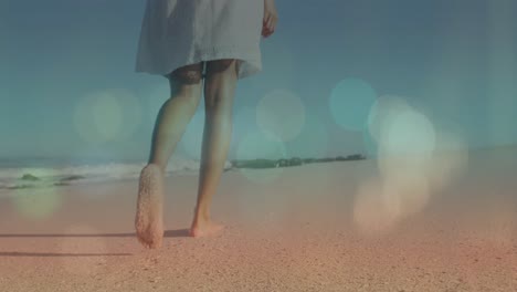 Animation-of-lens-flare-over-low-section-of-biracial-young-woman-walking-barefoot-on-sandy-beach