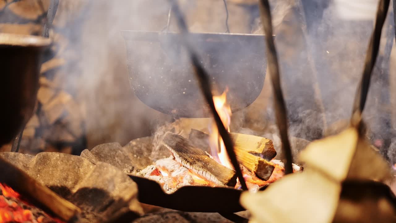 Slow motion of steaming cooking pot above glowing firewood with smoke rising