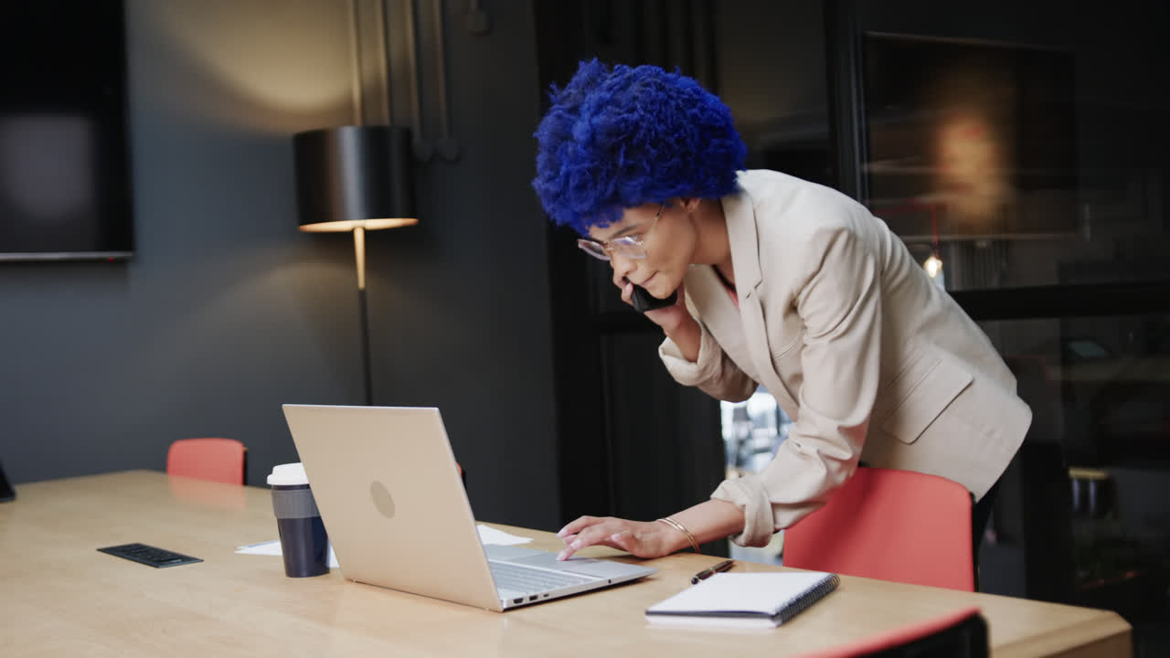 Biracial businesswoman with blue afro talking on smartphone and using laptop at desk, slow motion