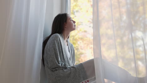 pensive woman sitting on a windowsill, looking outside through the window in the morning at home