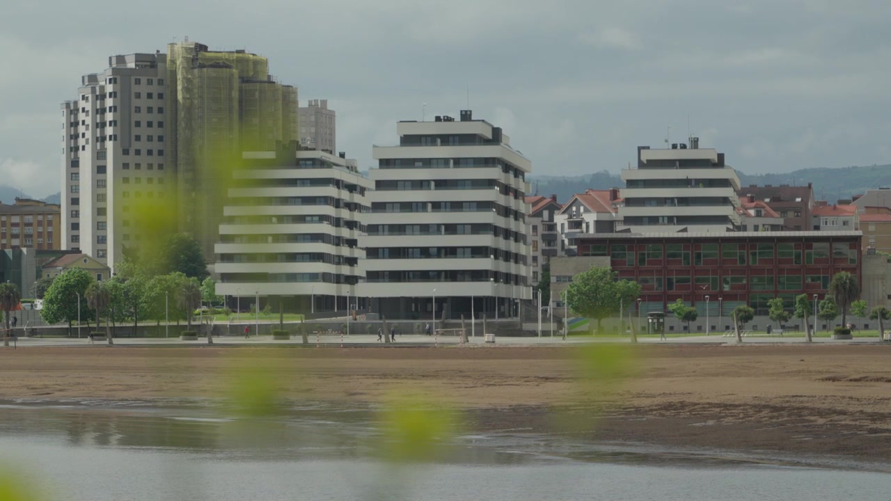 Toma de seguimiento diurna de modernos edificios residenciales escalonados a lo largo del paseo marítimo de la Playa del Arbeyal en Gijón, Asturias. Presenta un diseño de playa de arena bajo un