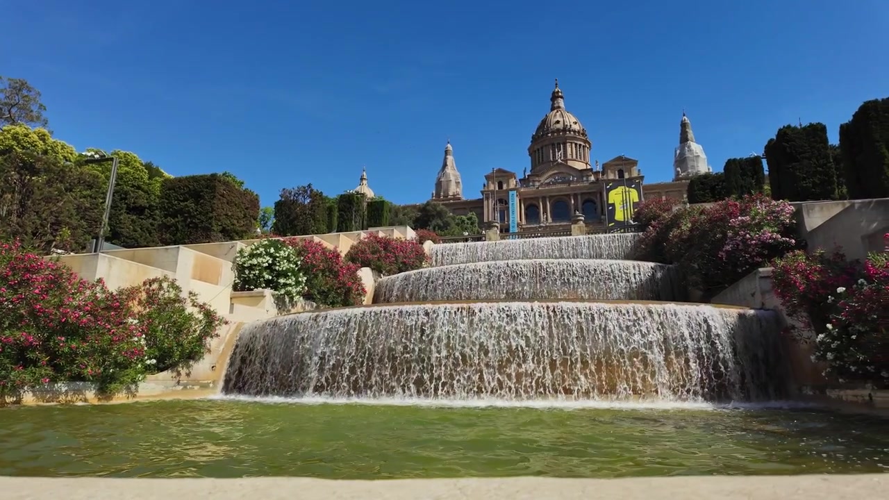 Plano deslizante dramático das fontes de água em cascata de Montjuïc em câmera lenta, com o grandioso Museu Nacional d'Art de Catalunya emoldurado por jardins vibrantes e céu azul claro.