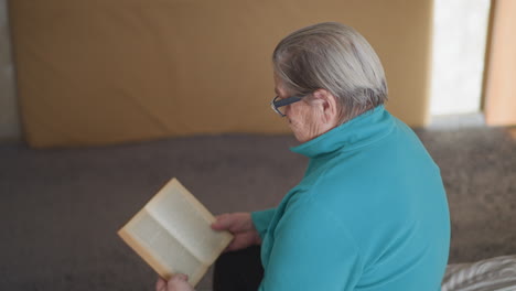 elderly pensioner in green teal sweater sitting on bed, reading book with focused attention. bright indoor lighting and peaceful setting, absorbed in reading with thoughtful expression