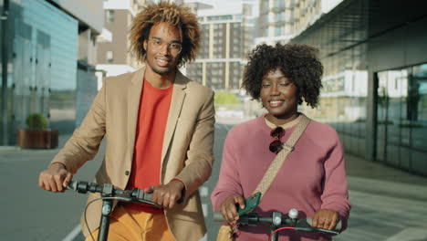 Portrait-of-Smiling-African-American-Man-and-Woman-with-E-Scooters-in-City
