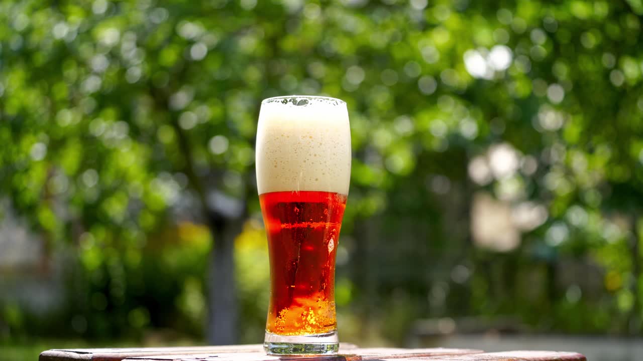 Cold beer in a glass with white foam. Craft beer in a glass with small bubbles inside. Cool brown beverage on a table outdoors. Close-up.