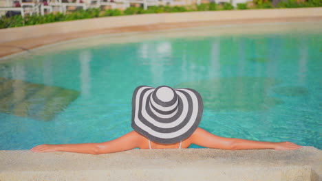 back of a woman in floppy striped hat leaning on concrete pool border inside swimming pool, no face template copy space