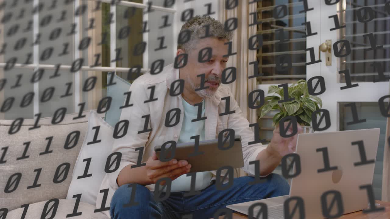 Mature man tapping laptop on sofa holding tablet, causing binary overlay and reviewing tech data