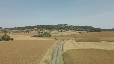 aerial images of tuscany in italy cultivated fields summer, flight over a dirt road with golden yellow cornfields beautiful little mountain town in the background