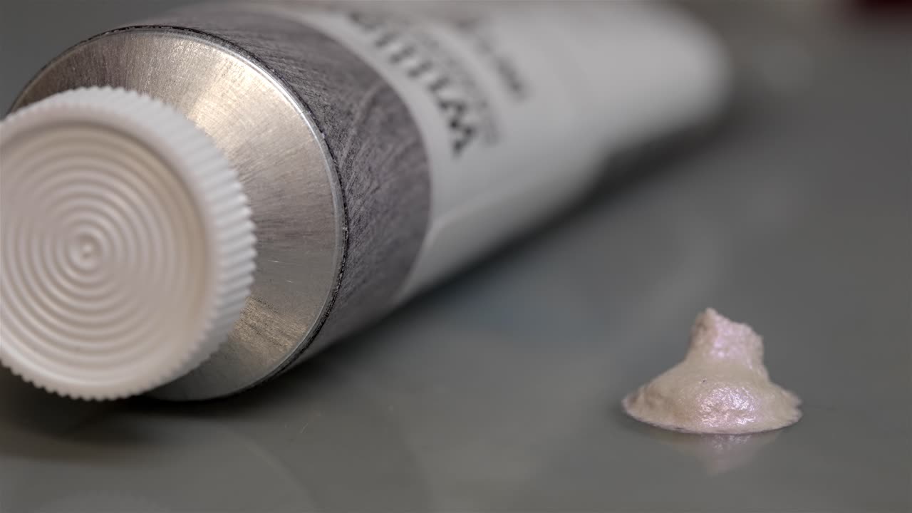 Shallow depth of field pan down across white tube of paint and glimmering pile