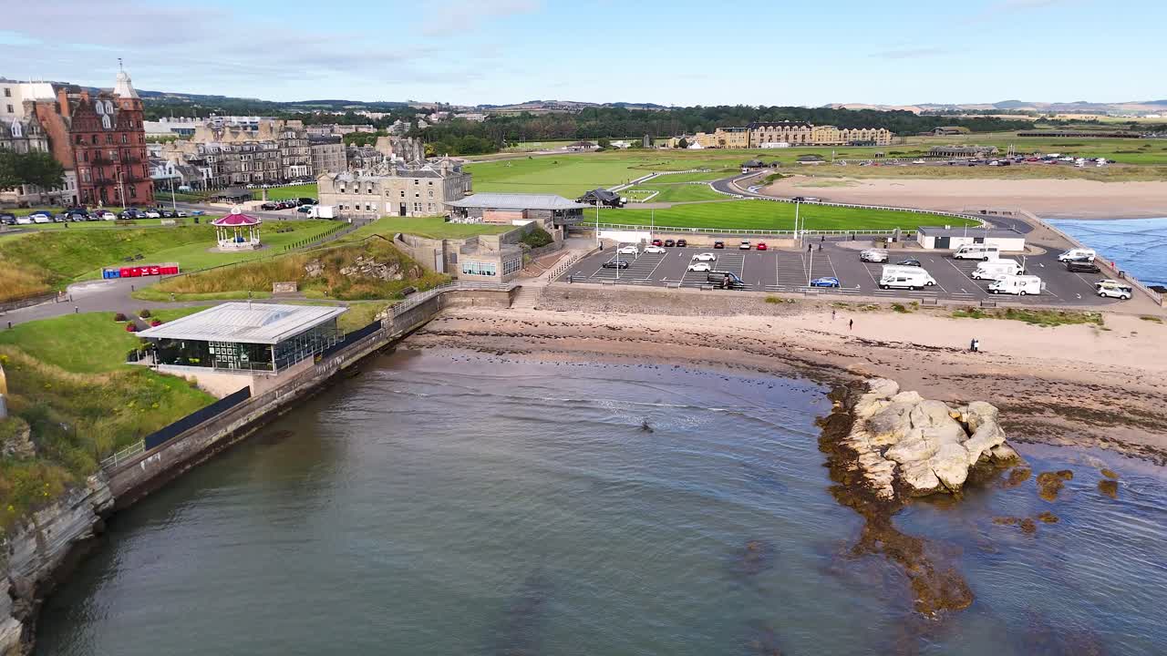 Drone glides above St Andrews waterfront, revealing Victorian architecture, rocky shoreline, parking area, and iconic golf course under bright daylight with steady movement