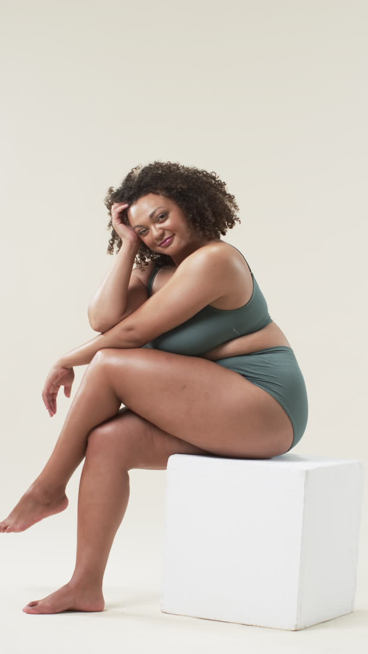 Vertical video: Sitting on white block, plus size woman in underwear embracing body positivity