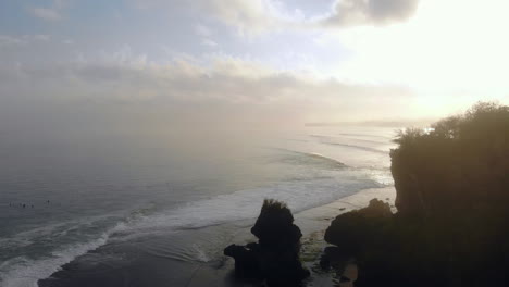 volando sobre el océano ondulante al amanecer, grupo de surfistas esperando la ola perfecta, aire brumoso, naturaleza y costa en el fondo