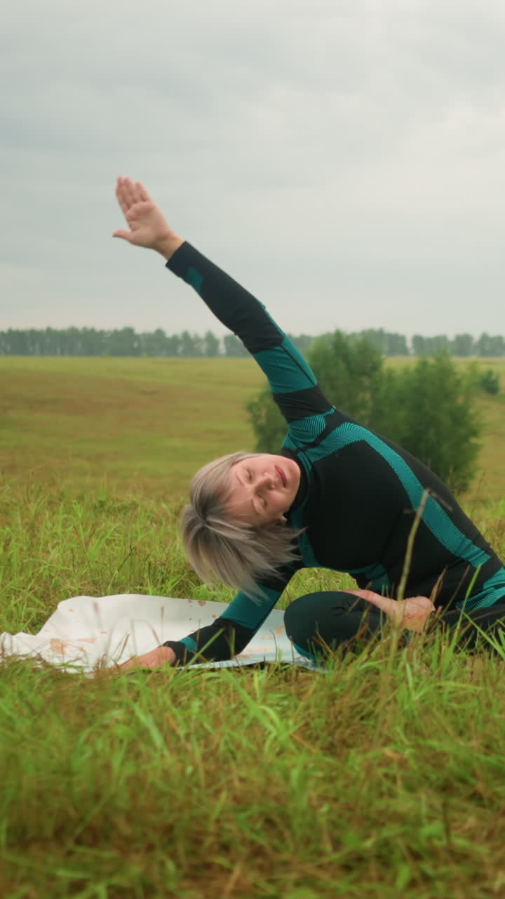 mujer avanzada acostada en un lado de la alfombra de yoga, practicando la postura de flexión lateral con el brazo extendido, sentada en un vasto campo de hierba bajo cielos nublados, rodeada de naturaleza y árboles en la distancia
