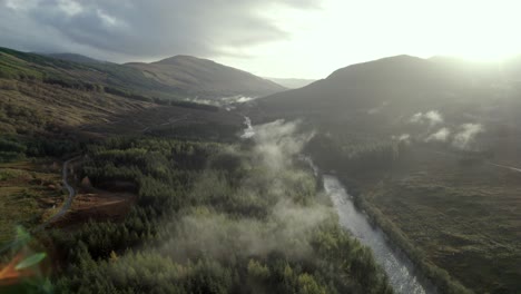 Imágenes-Aéreas-De-Drones-Volando-Alto-Y-Lentamente-Sobre-Un-Río,-Una-Carretera-Y-Un-Bosque-De-Coníferas-Mientras-Una-Nube-Baja-Abraza-Las-Copas-De-Los-árboles-Y-El-Sol-Se-Pone-Detrás-De-Las-Montañas