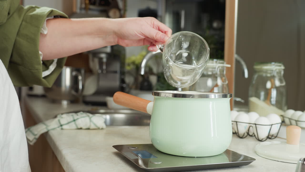 Clean water being poured from transparent glass into pastel green jar with wooden handle placed on digital kitchen scale, surrounded by eggs, jars, and utensils in cozy home kitchen
